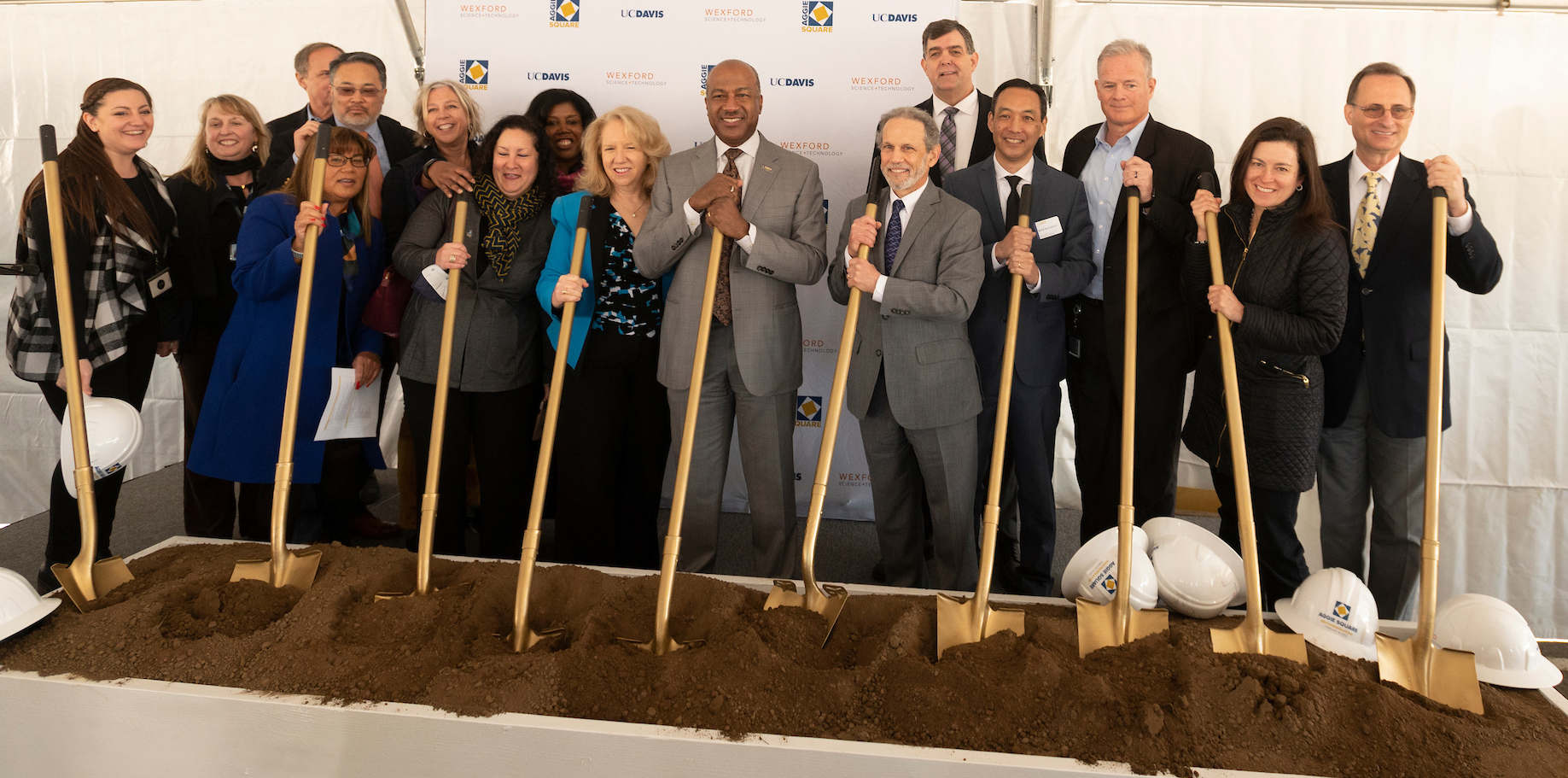 A group of people stand with shovels over a dirt trough.