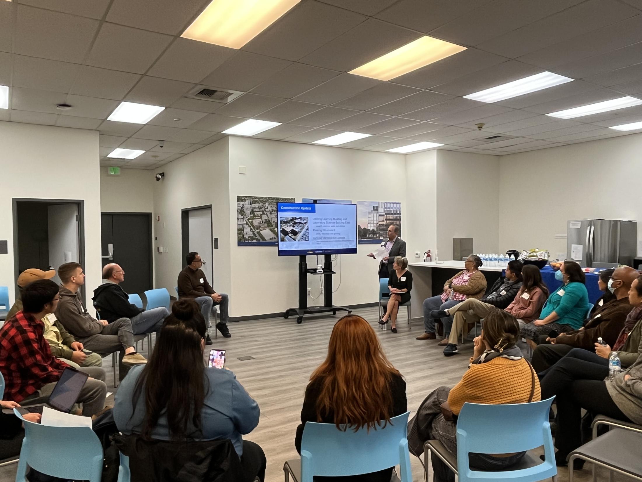 Image of UC Davis Aggie Square's quarterly community engagement meeting. People are sitting in a circle of chairs listening to a presenter in front of an LCD screen with slides.