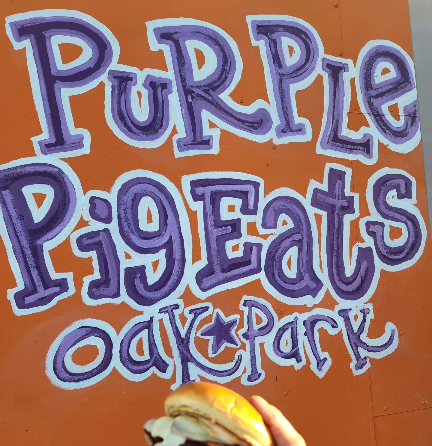 A burger in front of an orange sign with purple text that reads "Purple Pig Eats, Oak Park"