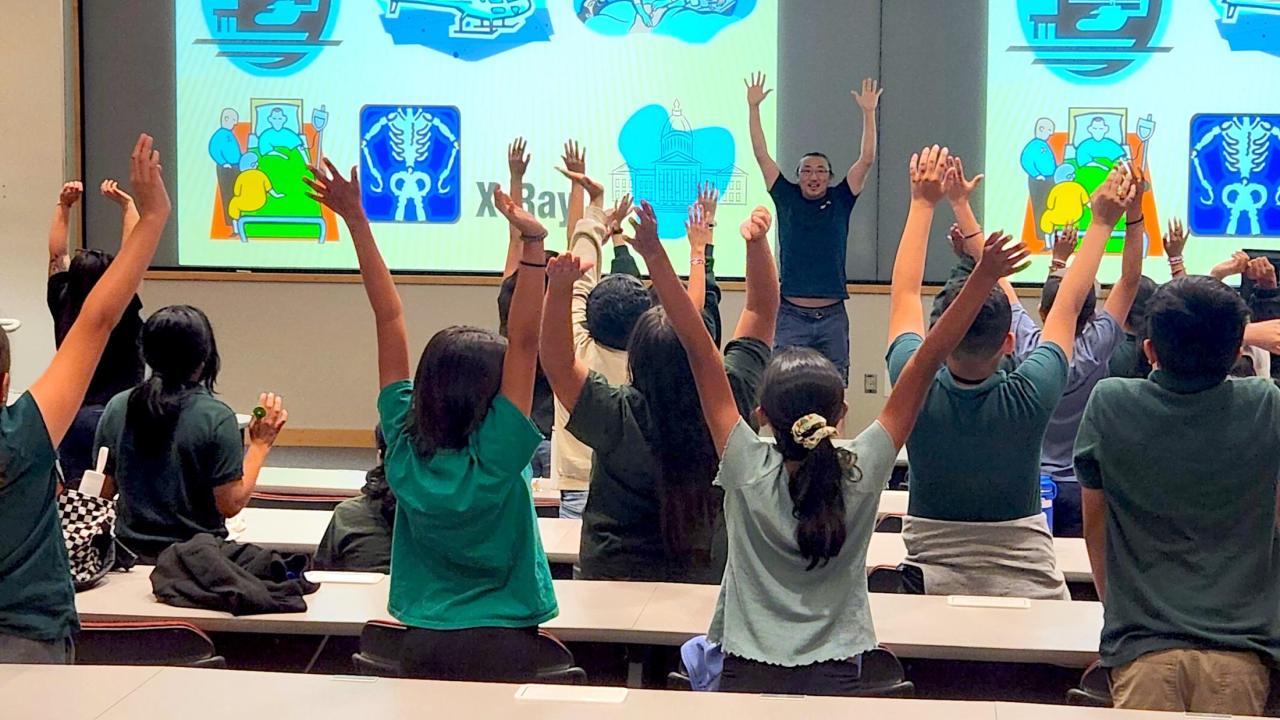Students in a lecture hall raise their hands above their heads.