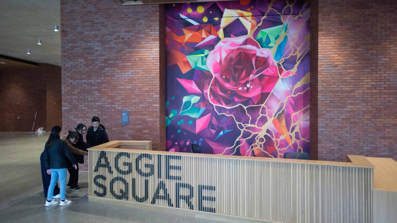 Students mingle next to the reception desk in front a brick wall with a large mural of a flower on it.