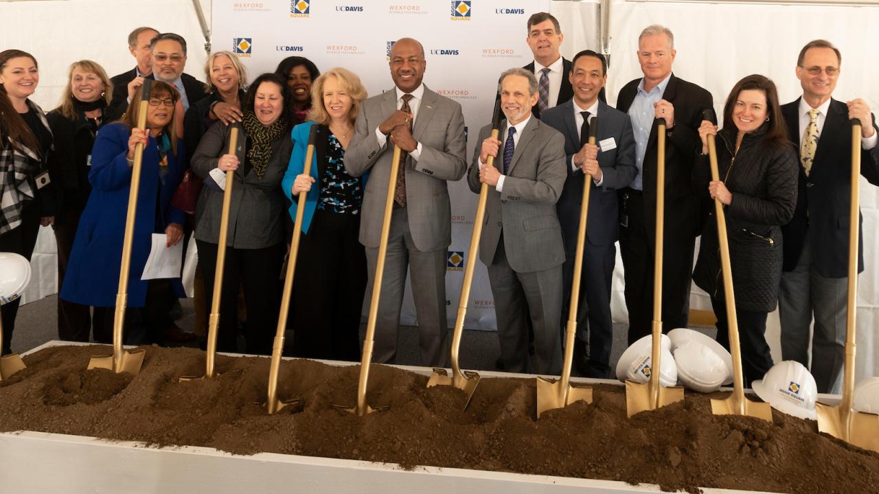A group of people stand with shovels over a dirt trough.