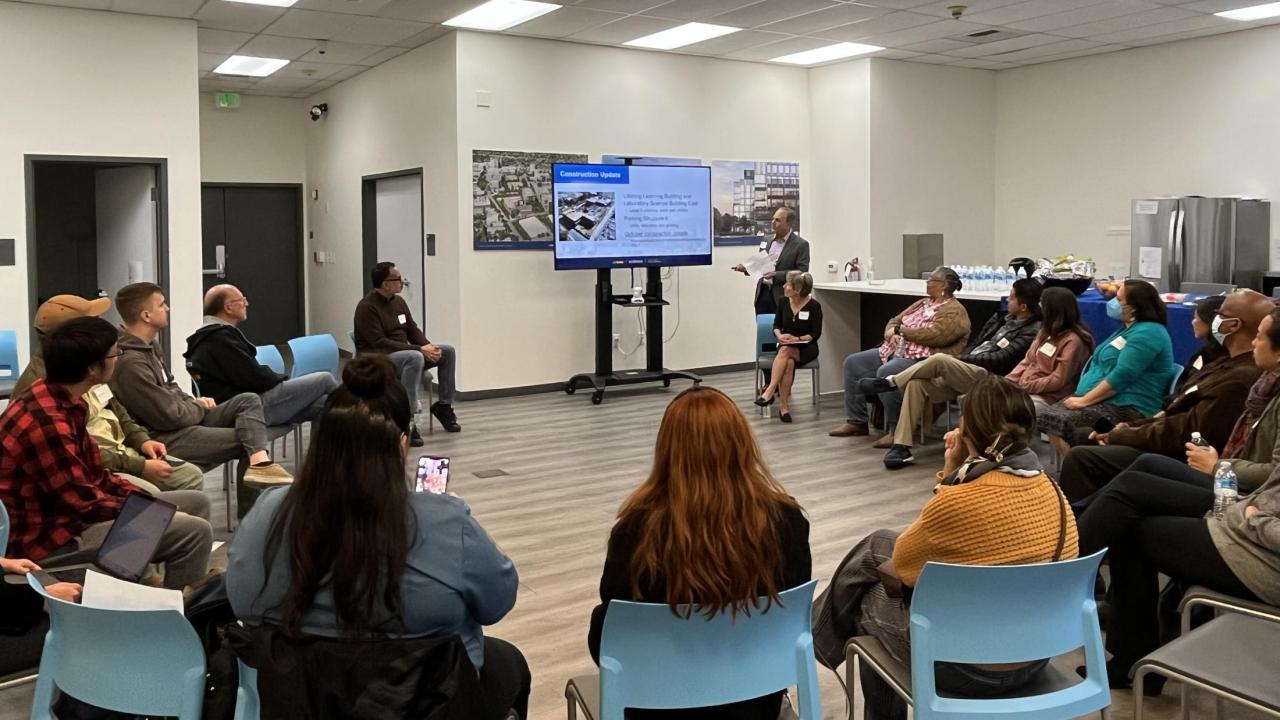 Image of UC Davis Aggie Square's quarterly community engagement meeting. People are sitting in a circle of chairs listening to a presenter in front of an LCD screen with slides.