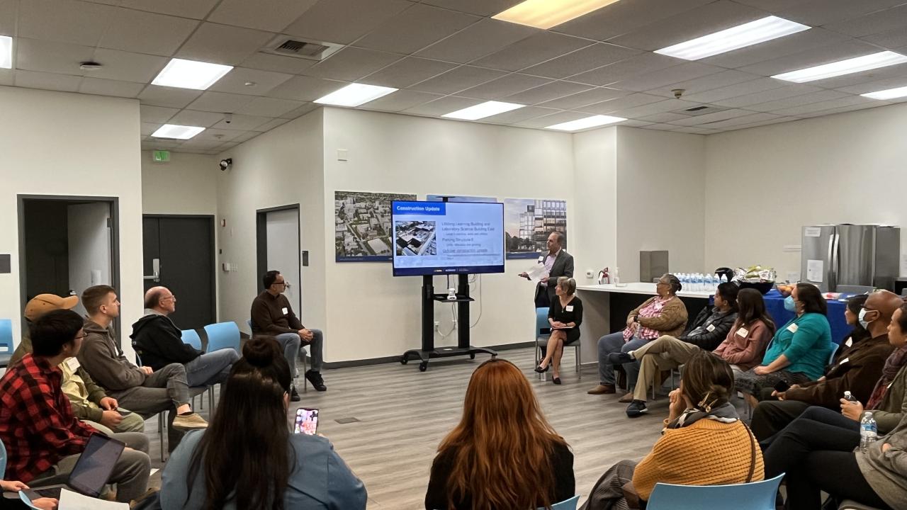 Image of UC Davis Aggie Square's quarterly community engagement meeting. People are sitting in a circle of chairs listening to a presenter in front of an LCD screen with slides.