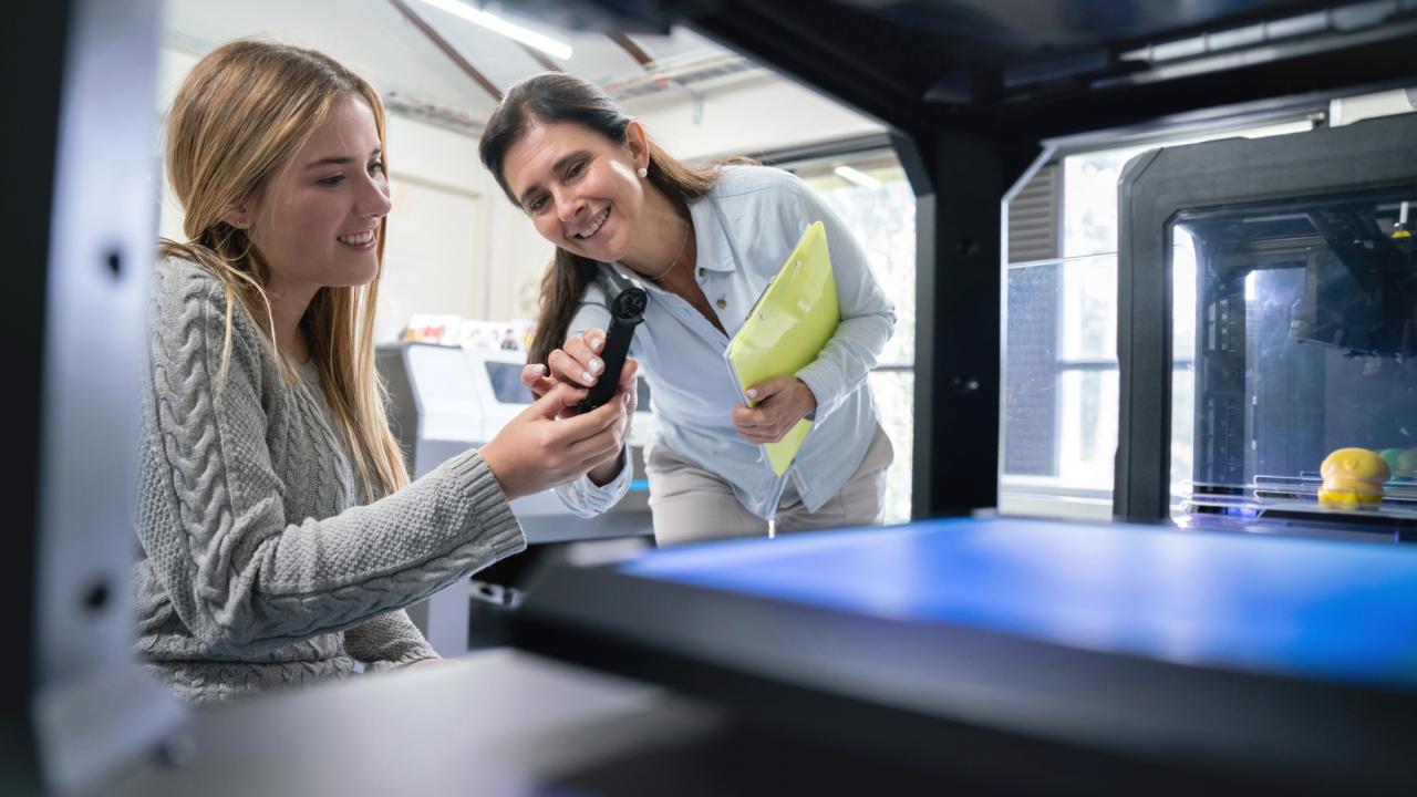 Two women look at an item that came from a big 3D printer