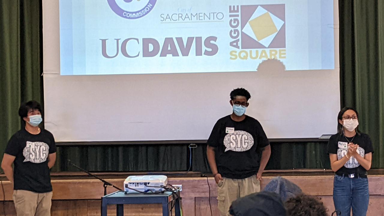Three students stand on a stage with a projection screen behind them.