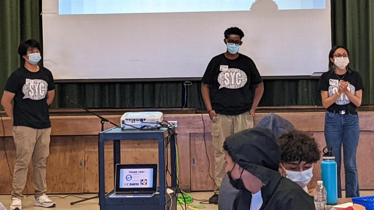 Three students stand on a stage with a projection screen behind them.