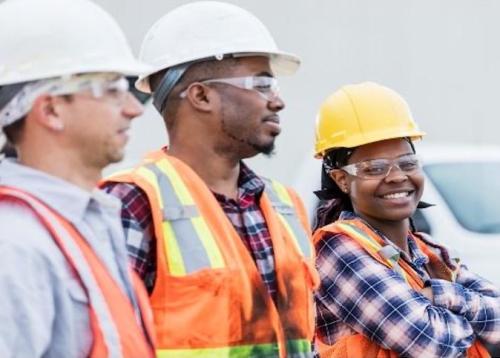 Image of three construction workers of diverse backgrounds. The female is looking at the camera and smiling.