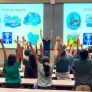 Students in a lecture hall raise their hands above their heads.