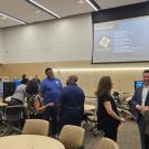 A large gathering of people talk in small groups in a room with a projector behind them.