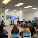 Image of UC Davis Aggie Square's quarterly community engagement meeting. People are sitting in a circle of chairs listening to a presenter in front of an LCD screen with slides.