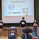 Three students stand on a stage with a projection screen behind them.